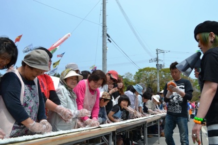 屋外に設置された長いテーブルの前に多くの参加者が横一列に並び、長い巻きずし作りを楽しんでいる様子を写した写真