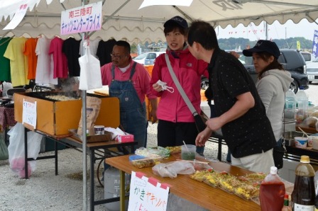 出店に「カキ焼きそば」がパック詰めして売られており、手前のテーブルに並べられた商品が見え、奥ではスタッフが鉄板で調理をしている様子を写した写真