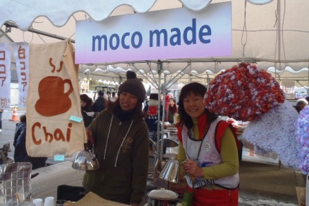 屋外のテントに「moco made」の看板が掲げられ、チャイを販売している二人の女性が笑顔でドリップポットを手にし写っている写真