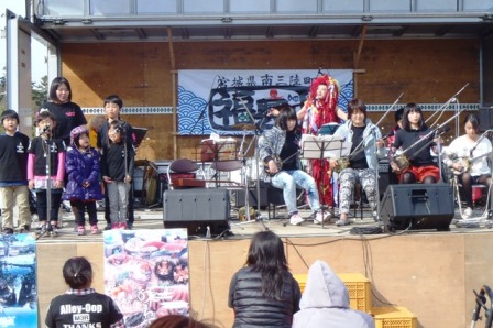 ステージ上で三味線のような楽器を演奏するグループと、その横に立つ子どもたちの合唱隊が並び、観客が見守る中で和やかに演奏が行われている様子を写した写真