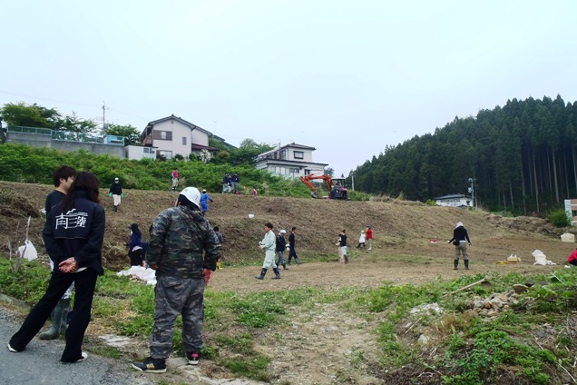 住宅地の下の斜面で多くの人々が農作業に取り組んでいる様子を写した写真