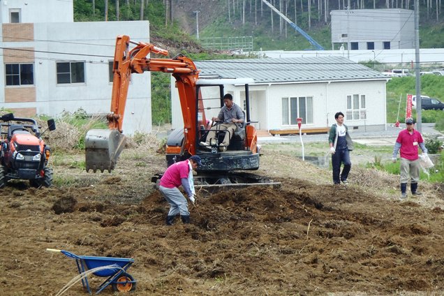 小型ショベルカーで耕されている畑の中で、前方の人が鍬で土を掘り起こしている様子を写した写真