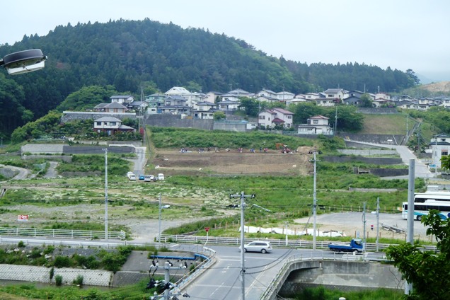 高台から撮影された住宅街と畑の全景で、中央の茶色い畑では人々が作業しており、手前には道路と車が見える写真