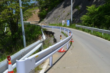 海沿いに伸びる道路の一部が崩れ、その手前にガードレールが設置されて細くなった道路の写真