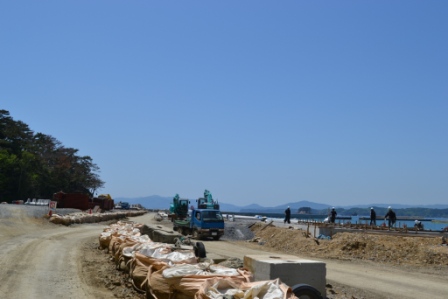 海岸沿いに重機が数台並び、作業員がヘルメットを着用して舗装や整備作業を行っている様子の写真