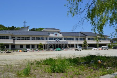 広々とした校庭の奥に白い外壁の二階建ての学校校舎が建ち、その周囲には工事用の資材や機材が置かれた工事中の名足小学校の写真