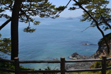 木々と木製の柵が並び、青い海が広がり遠くに浮かぶ島々や連なった山々を歌津崎から撮影した写真