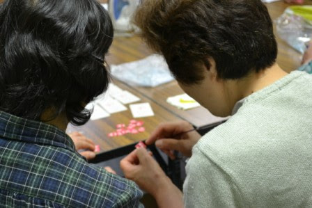 2人の女性が小さな布やパーツを使って手作業で細かい作業をしている様子を背後から写した写真