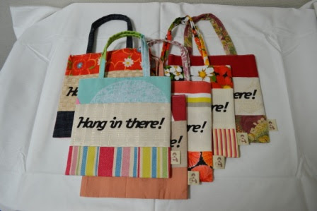 「Hang in there!」というメッセージが書かれた色とりどりの布製トートバッグが6つ、丁寧に並べられている写真