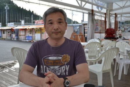 屋外の飲食スペースで白い椅子とテーブルに囲まれて、南三陸ミシン工房代表の熊谷安利さんがカメラの前で手を組んで座っている写真