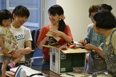 若い女性が箱の中から布や材料を取り出して見せており、まわりの女性や子どもが笑顔で囲んでいる写真