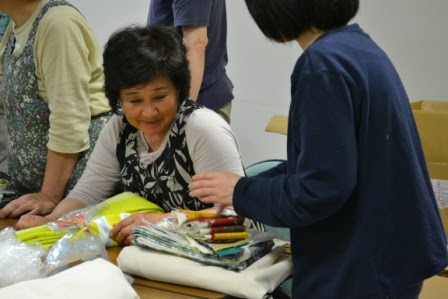 テーブルの上に並んだ布や糸を前に、エプロンをした女性が笑顔で座り、別の女性と会話している写真