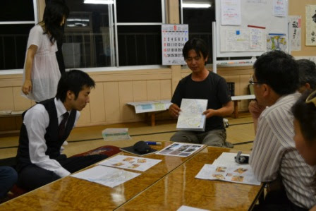 白い紙を持った男性が紙を指差しながら発表しており、周りの人が興味深く話を聞いている様子の写真