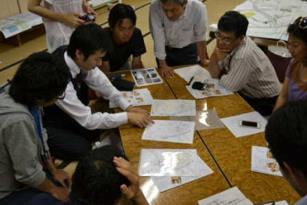 複数の人が室内で一つのテーブルを囲み、地図や紙の資料を広げながら互いに意見を交わしている様子が写っている写真