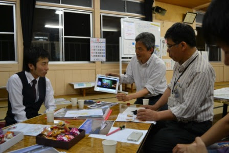 テーブルを囲んで座っており、1人の男性がタブレットを持ち上げて他の参加者に画面を見せている様子が写っている写真