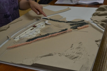 机の上に置かれた地形の立体模型を一人の人物が指差して説明している様子が写っている写真