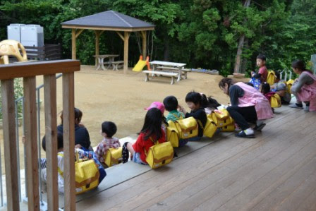 園舎のウッドデッキで黄色い通園バッグを背負った幼児たちが横並びに座っており、目の前には東屋や木製のテーブルとイスが置かれている園庭が広がっている様子の写真