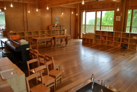 木の床と天井に囲まれた広い室内に、子ども用の机と椅子、絵本棚やピアノが配置され、窓から自然光が差し込む園内の写真