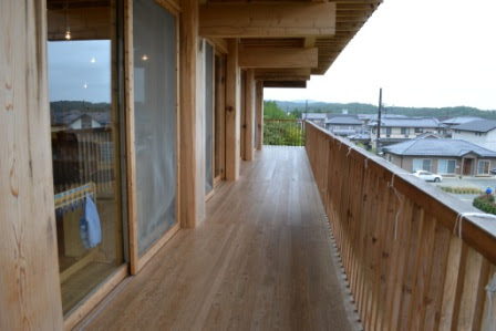 園舎の長い木製の回廊が建物に沿って続き、右手に住宅街が広がる景色が見える写真