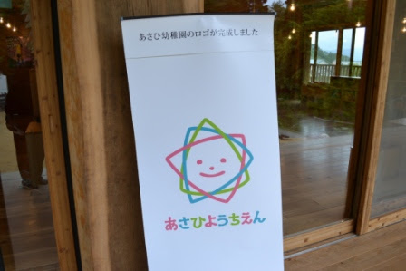 「あさひ幼稚園のロゴが完成しました」と書かれた案内板に、カラフルな線で囲まれた笑顔の顔と「あさひようちえん」という文字が描かれている写真