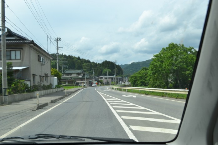 曇り空が広がる中、右にカーブした道路とその周囲の住宅、電柱、山々、木々が広がる風景を、車のフロントガラス越しに見た写真