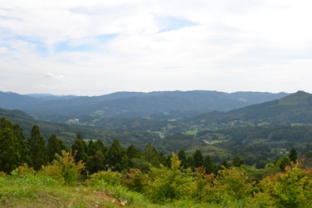 澄んだ青空が広がる、背景に連なる丘や山々に囲まれ、緑豊かな草木が広がる盆地に家が点在する、美しい風景を高台から撮影した写真