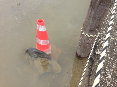 赤白のカラーコーンが濁った水に半分沈んでおり、隣の電柱にはロープが巻かれている写真