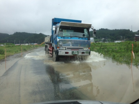 大きな青いダンプトラックが水のたまった道路を通過し、水をはね上げている写真