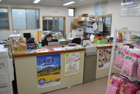 カウンターの奥で職員が仕事をしており、棚には書類や事務用品、手前には農業関連のチラシや商品が並ぶJAの事務所内の様子を写した写真