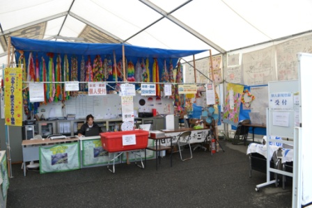 白いテントの中で、折り鶴や子どもたちの絵などが飾られたカラフルな装飾の下に複数の机と椅子が並び、受付のような雰囲気の災害ボランティアセンター内部の写真