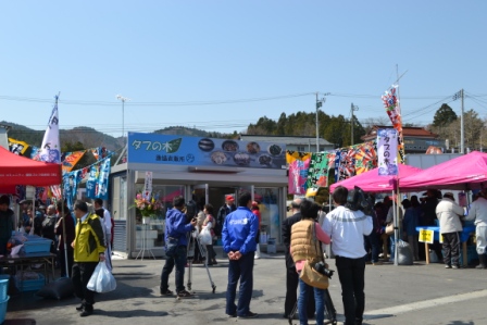 「タブの木」と書かれた看板が掲げられた漁協の直売所の前に、地元の人々や観光客が集まり、カラフルなのぼり旗やテントが立ち並ぶ中で、買い物や会話を楽しみながらにぎやかに交流している様子の写真