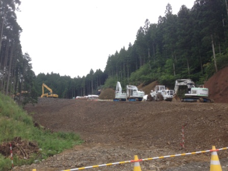 森林の中を切り開いて造成された道路予定地に、複数の重機が並んで待機している工事現場を写した写真