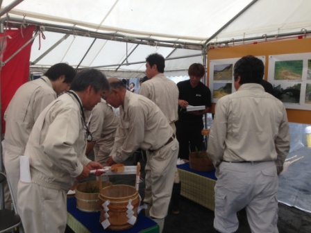 安全祈願祭のために設置されたテント内で、作業服姿の関係者たちが供え物の準備をしている、儀式の直前の様子の写真