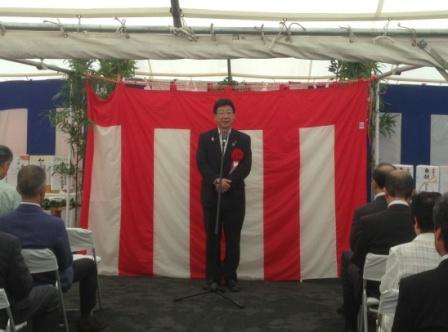 赤白の幕の前に立つ黒いスーツ姿の佐藤仁町長が、マイクを持って挨拶を行っている、安全祈願祭での来賓挨拶の写真