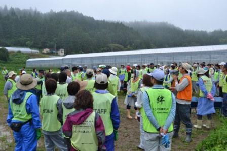 周囲は曇り空で山に囲まれた農村風景が広がっている田畑の前で多数のボランティアが緑色のビブスを着て整列し、スタッフの説明を聞いている様子の写真