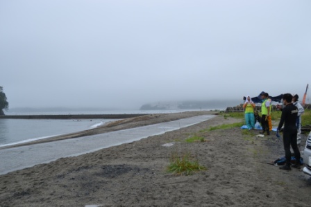 砂浜と海に向かって数名が立ち、遠くには防潮堤と海が広がる中、活動を見守るスタッフの姿もある、清掃活動の全体情景を捉えた写真