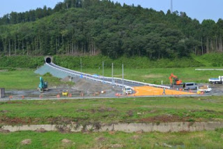 緑に囲まれた丘のふもとにある工事現場で、道が右から丘の左下のトンネルへと続き、複数のショベルカーやトラックなどの建設車両が作業をしている様子が写された、開発中の風景の写真