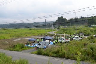 曇り空の下で、遠くの丘が広がる手前に道路が通り、複数のテントと車両が並ぶ広々とした野原に、蛍光色のベストを着た人々が集まって活動している写真