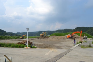 空には雲が広がり、舗装されたエリアと建設資材が点在する緑の丘と木々に囲まれた場所で、三台のショベルカーが大きな土の山を掘削している工事現場の写真