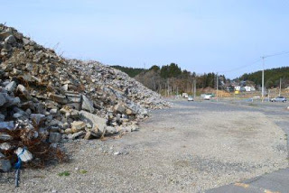 木々と建物が見える風景で、右側には砂利が広がる空間が続き、左側に大きな瓦礫の山がある、解体後の様子を写した写真