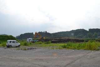 緑の丘と木々が広がる舗装された空間の手前に雑草が生い茂り、左側には白いバンが停車していて、奥にはショベルカーとトラックが作業している工事現場の写真