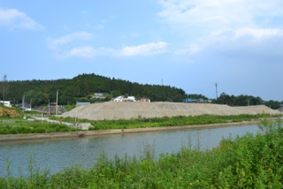青空と雲が広がる奥には丘の木々と点在する建物が見え、手前に川が流れ、その両岸には緑の草木が茂り、中ほどには砂や砂利の大きな山がある風景の写真