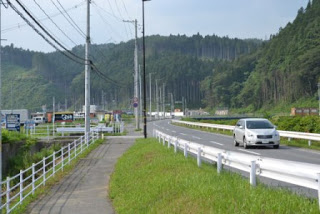 背景には緑の草と木々に囲まれ、右方向に緩やかにカーブしている道路を白い車が走っており、左側には白いガードレールが設置された歩道がある写