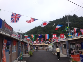 通りの上にさまざまな国旗がひもで吊るされ、小さな店舗や屋台が並ぶ道を人々が歩いている、賑やかなさんさん商店街の写真