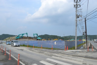 背景に丘と曇り空が広がる、車が走行している道路が交差する角地に、青と白のフェンスに囲まれた工事現場で複数の重機が作業している写真