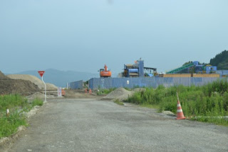 周囲には緑と遠くの丘が見え、砂利道の先にオレンジ色の重機や土の山が並ぶ、青いフェンスで囲まれた工事現場の写真