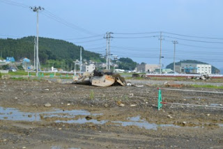 青空の下、奥には建物や森のある丘が見える中央に、水たまりとがれきがある泥だらけの空き地が広がる写真