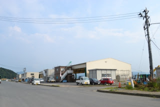 青空の下、左奥には山が見える手前に建つ、周囲に車両が駐車している仮設の志津川魚市場の写真