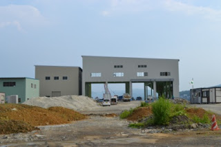 青空の下、土の山や資材のある奥に、工事車両が並ぶ大きな窓や開口部のある建設中の造船所の写真