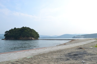 遠くの山々が広がり、砂浜の先にコンクリートの桟橋が伸び、穏やかな海に浮かぶ小さな森の島の写真